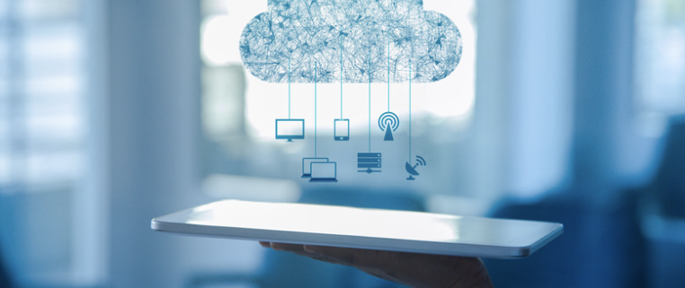 A hand holding a tablet with a holographic cloud icon above it. The cloud icon is interconnected with various digital devices, representing the integration of cloud infrastructure services. The background is a blurred office setting, highlighting the professional and business context of cloud computing.