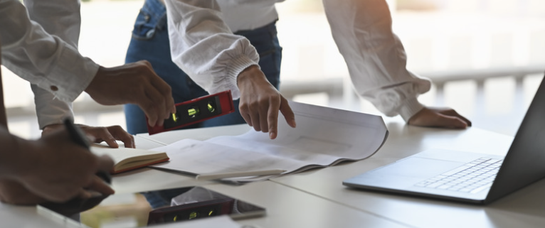 Two professionals collaborating at a desk, examining blueprints and documents. One person is using a level tool to ensure accuracy. The scene also includes a laptop and tablet, highlighting the integration of technology in planning and decision-making processes. The image represents the importance of meticulous planning and infrastructure management in business continuity.